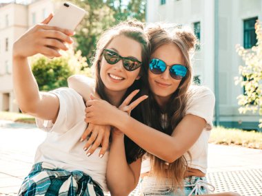 Son moda beyaz tişörtlü iki güzel gülümseyen hippi kadının portresi. Sokakta poz veren kaygısız kadınlar. Pozitif modeller eğleniyor, sarılıyor ve selfie çekiyor.