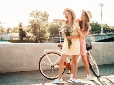 İki genç, güzel, gülümseyen yeniyetme kadın, modaya uygun bir yaz elbisesi içinde. Kaygısız kadınlar geçmişe dönük bisiklete biniyor. Pozitif modeller bisiklette poz verirken eğleniyorlar.