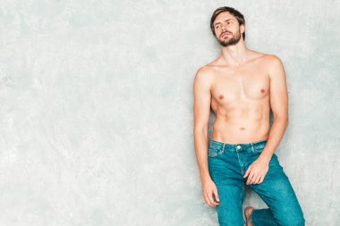 Yakışıklı, güçlü bir adamın portresi. Kot pantolonla gri duvarın yanında poz veren sağlıklı gülümseyen atletik form modeli. Çıplak gövdesi olan kendine güvenen seksi moda erkeği. Lambersexual stüdyoda.
