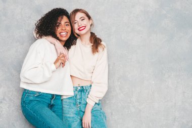 İki güzel, güzel, gülümseyen uluslararası hippi kadın, son moda yaz kot kıyafetleri içinde. Stüdyoda gri duvarın yanında poz veren kaygısız seksi kadınlar. Pozitif modeller eğleniyor. Arkadaşlık kavramı 