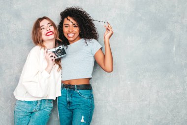 İki güzel, güzel, gülümseyen uluslararası hippi kadın, son moda yaz kot kıyafetleri içinde. Stüdyoda gri duvarın yanında poz veren kaygısız seksi kadınlar. Pozitif modeller eğleniyor. Retro kamerada fotoğraf çekmek