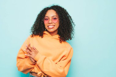 Afro kıvırcık saçlı, turuncu kapüşonlu ve kotlu güzel siyah kadın. Stüdyoda mavi duvarın yanında poz veren kaygısız seksi kadın. Güneş gözlükleriyle bronzlaşmış ve neşeli