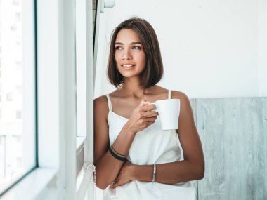 Beyaz pijama giymiş güzel bir kadının portresi. Kaygısız kadın elinde fincanla kahve içiyor. Seksi manken sabah kahvaltısını balkonda yapıyor. Hayal kurar