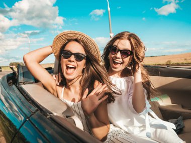Üstü açık arabadaki iki güzel ve gülümseyen hippi kadının portresi. Cabriolet kullanan kaygısız seksi kadınlar. Pozitif modeller güneş gözlükleriyle geziyor ve eğleniyorlar.