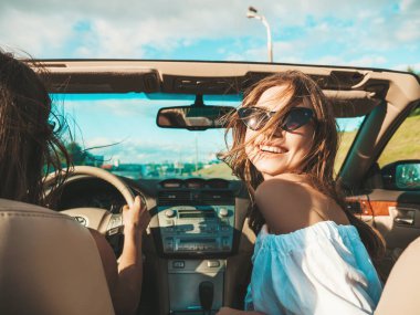 Üstü açık arabadaki iki güzel ve gülümseyen hippi kadının portresi. Cabriolet kullanan kaygısız seksi kadınlar. Pozitif modeller güneş gözlükleriyle geziyor ve eğleniyorlar.
