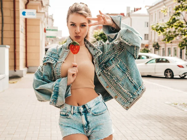 Hipster kot ceketli güzel bir mankenin portresi. Lolipop karpuz şekeri yiyen bir kadın. Sokakta poz veren bir kadın. Dışarıdaki kadın.