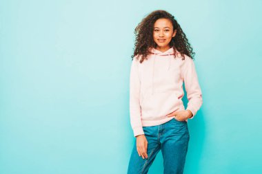 Afro kıvırcık saçlı güzel siyah bir kadın. Pembe kapüşonlu ve şık kot elbiseli bir model. Stüdyoda gri arka planda poz veren kaygısız seksi kadın. Bronzlaşmış ve neşeli