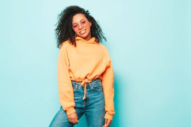 Afro kıvırcık saçlı, turuncu kapüşonlu ve kotlu güzel siyah kadın. Stüdyoda mavi duvarın yanında poz veren kaygısız seksi kadın. Güneş gözlükleriyle bronzlaşmış ve neşeli