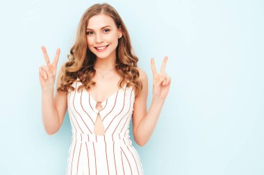 Son moda yaz hippi kıyafetleri içinde gülümseyen güzel bir kadının portresi. Stüdyoda açık mavi duvarın yanında poz veren kaygısız seksi kadın. Pozitif model içeride eğleniyor. Barış işareti gösterir