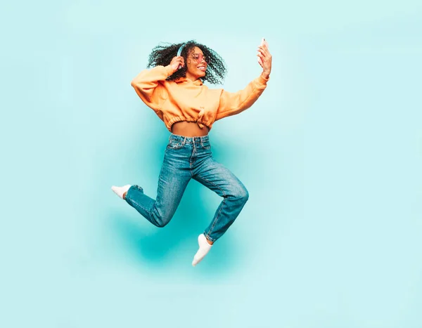 Beautiful black woman with afro curls hairstyle.Smiling model orange hoodie.Sexy carefree female listening music in wireless headphones. Jumping in studio on blue background.Dynamic movement