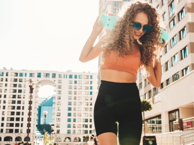 Saçları kıvırcık, genç, güzel, gülümseyen hippi kadın. Yazlık giysiler içinde son moda bir model. Sokakta mavi paralı kaykaylı kadın poz veriyor.