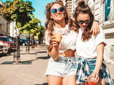 Moda kıyafetleri içinde gülümseyen iki güzel hippi kadın. Dışarıda poz veren kaygısız kadınlar. Pozitif modeller, plastik bardakta pipetle taze kokteyl içiyorlar ve içiyorlar.