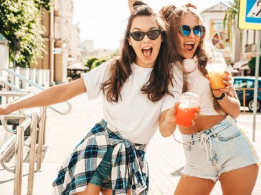 Moda kıyafetleri içinde gülümseyen iki güzel hippi kadın. Dışarıda poz veren kaygısız kadınlar. Pozitif modeller, plastik bardakta pipetle taze kokteyl içiyorlar ve içiyorlar.