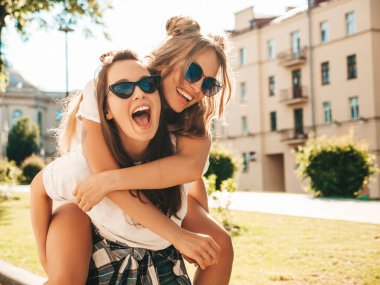 Son moda beyaz tişörtlü iki güzel, gülümseyen yeniyetme kadın sokak arka planında poz veren kaygısız kadınlar. Manken arkadaşının sırtına atlayıp, onu dışarı taşır.