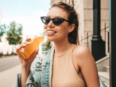 Yazlık hippi kot ceketli güzel bir mankenin yakın plan portresi. Sokakta poz veren kıyafetler. Güneş gözlüklü bir şişeden renkli bir kokteyl içmek. Gülümseyen moda kadını.