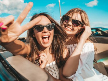 Üstü açık arabadaki iki güzel ve gülümseyen hippi kadının portresi. Cabriolet kullanan kaygısız seksi kadınlar. Pozitif modeller dışarıda güneş gözlüğü takıp eğleniyorlar. Yaz günlerinin tadını çıkarıyorlar.