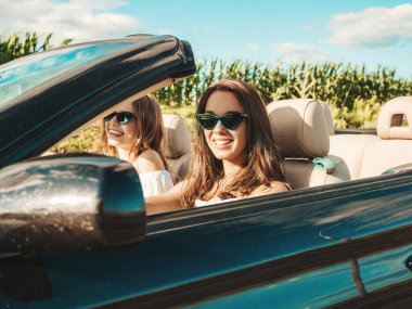 Üstü açık arabadaki iki güzel ve gülümseyen hippi kadının portresi. Cabriolet kullanan kaygısız seksi kadınlar. Pozitif modeller dışarıda güneş gözlüğü takıp eğleniyorlar. Yaz günlerinin tadını çıkarıyorlar.