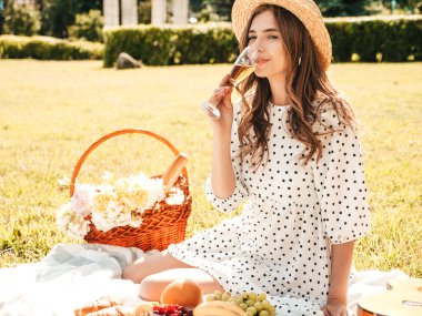 Moda elbisesi ve şapkası olan genç ve güzel hippi kadın. Kaygısız kadın dışarıda piknik yapıyor. Pozitif model çimlerin üzerinde oturuyor, meyve ve peynir yiyor, şampanya içiyor.