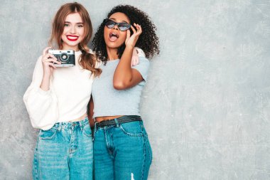 İki güzel, güzel, gülümseyen uluslararası hippi kadın, son moda yaz kot kıyafetleri içinde. Stüdyoda gri duvarın yanında poz veren kaygısız seksi kadınlar. Pozitif modeller eğleniyor. Retro kamerada fotoğraf çekmek