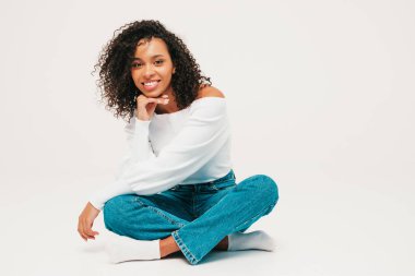 Saçları kıvırcık, güzel siyah bir kadın. Süveter ve kotlar içinde gülümseyen bir model. Stüdyoda beyaz arka planda oturan kaygısız seksi kadın. Bronzlaşmış ve neşeli