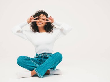 Saçları kıvırcık, güzel siyah bir kadın. Süveter ve kotlar içinde gülümseyen bir model. Stüdyoda beyaz arka planda oturan kaygısız seksi kadın. Bronzlaşmış ve neşeli. Barış işareti gösterir