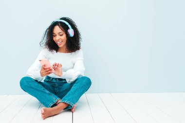 Saçları kıvırcık, saçları kıvırcık, süveter ve kot giyen, kablosuz kulaklıkla müzik dinleyen kaygısız bir kadın, açık mavi duvarın yanındaki stüdyoda. Akıllı telefonu tutar