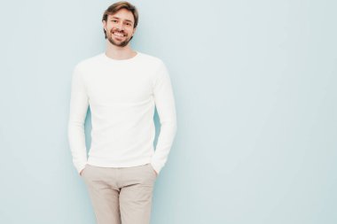 Beyaz süveter ve pantolon giyen yakışıklı, gülümseyen hippi, keresteci iş adamı modeli portresi. Moda tarzı bir adam stüdyoda açık mavi duvara karşı poz veriyor.