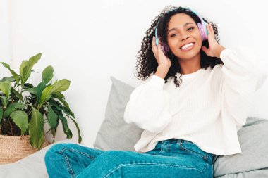 Saçları kıvırcık, saçları kıvırcık, süveter ve kot giyen güzel siyah bir kadın sabahları kablosuz kulaklıkla müzik dinleyen kaygısız bir kadın.