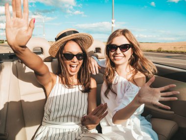 Üstü açık arabadaki iki güzel ve gülümseyen hippi kadının portresi. Cabriolet kullanan kaygısız seksi kadınlar. Pozitif modeller dışarıda güneş gözlüğü takıp eğleniyorlar. Yaz günlerinin tadını çıkarıyorlar.