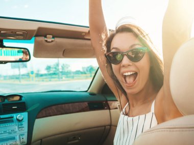 Üstü açık arabadaki güzel ve gülümseyen yeniyetme kadın portresi. Cabriolet kullanan kaygısız seksi kadın. Pozitif model sürüş ve gün batımında güneş gözlüğüyle eğlenme. El kaldırma.