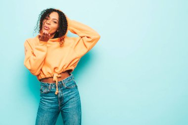 Afro kıvırcık saçlı, turuncu kapüşonlu ve kotlu güzel siyah kadın. Stüdyoda güneş gözlükleriyle mavi duvarın yanında poz veren kaygısız seksi kadın. Hava öpücüğü verir