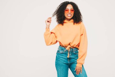 Afro kıvırcık saçlı, turuncu kapüşonlu ve kotlu güzel siyah kadın. Stüdyoda beyaz arka planda poz veren kaygısız seksi kadın. Güneş gözlükleriyle bronzlaşmış ve neşeli