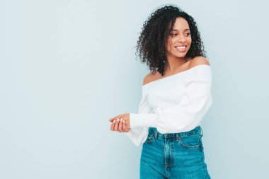 Saçları kıvırcık, güzel siyah bir kadın. Süveter ve kotlar içinde gülümseyen bir model. Stüdyoda açık mavi duvarın yanında poz veren kaygısız seksi kadın. Bronzlaşmış ve neşeli