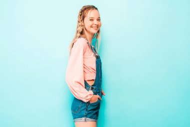 Genç ve güzel bir kadın kameraya bakıyor. Popüler bir bayan günlük kot pantolon içinde gülümsüyor. Stüdyoda mavi duvarın yanında poz veren pozitif ve komik bir model. Neşeli ve mutlu 