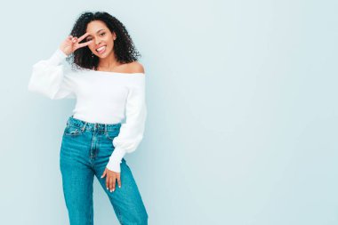 Saçları kıvırcık, güzel siyah bir kadın. Süveter ve kotlar içinde gülümseyen bir model. Stüdyoda açık mavi duvarın yanında poz veren kaygısız seksi kadın. Bronzlaşmış ve neşeli. Barış işareti nasıl?