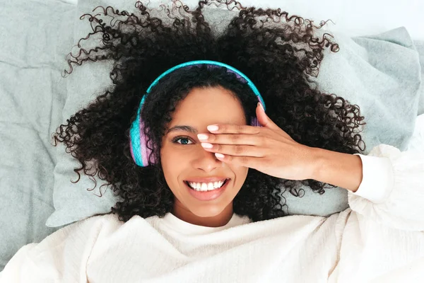 Beautiful black woman with afro curls hairstyle.Smiling model in sweater and jeans.Sexy carefree female listening music in wireless headphones in the morning.Lying in bed in white interior. Top view