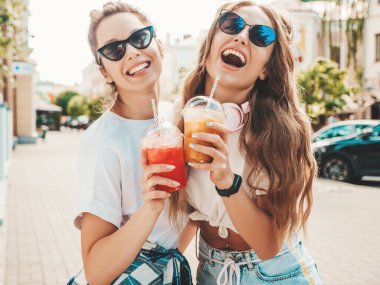 Moda kıyafetleri içinde gülümseyen iki güzel hippi kadın. Dışarıda poz veren kaygısız kadınlar. Pozitif modeller, plastik bardakta pipetle taze kokteyl içiyorlar ve içiyorlar.