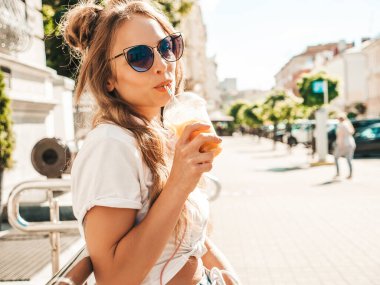 Genç, güzel, gülümseyen, hippi bir kadın. Yazın moda kıyafetleri içinde, sokakta poz veren bir kadın. Modeli elinde tutuyor ve samanla plastik bardakta taze sebze kokteyli içiyor.
