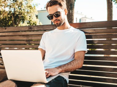 Yakışıklı, güler yüzlü, stil sahibi hipster Lambersex modeli. Moda adamı sokakta bankta oturuyor ve dizüstü bilgisayar kullanıyor, mesaj yazıyor. Ortak çalışma konsepti. Teknoloji ve iletişim