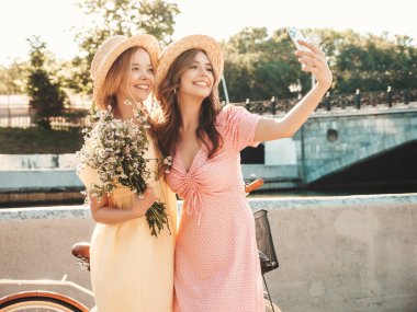 İki genç, güzel, gülümseyen yeniyetme kadın, modaya uygun bir yaz elbisesi içinde. Sokakta şapkalarla poz veren kaygısız seksi kadınlar. Sunset 'te pozitif modeller var. Selfii alıyorum. Çiçekleri tutuyorum.