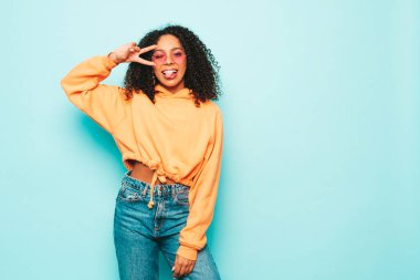 Afro kıvırcık saçlı, turuncu kapüşonlu ve kotlu güzel siyah kadın. Stüdyoda güneş gözlükleriyle mavi duvarın yanında poz veren kaygısız seksi kadın. Barış işareti gösterir