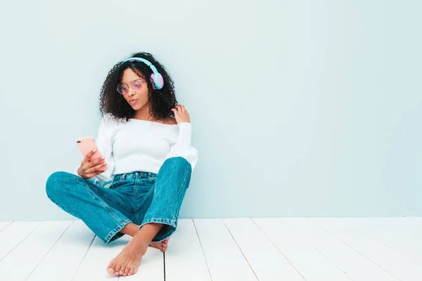 Beautiful black woman with afro curls hairstyle.Smiling model in sweater and jeans.Sexy carefree female listening music in wireless headphones.Sitting in studio near light blue wall. Holds smartphone