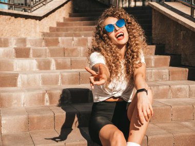 Kıvırcık saçlı güzel bir manken yaz hippi kıyafetleri giyiyor. Sokakta merdivenlerde oturan kaygısız bir kadın. Moda, komik ve olumlu kadın barış işareti gösteriyor.