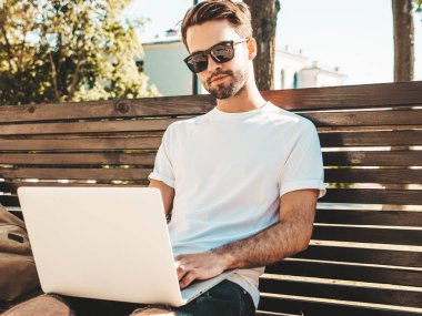 Yakışıklı, güler yüzlü, stil sahibi hipster Lambersex modeli. Moda adamı sokakta bankta oturuyor ve dizüstü bilgisayar kullanıyor, mesaj yazıyor. Ortak çalışma konsepti. Teknoloji ve iletişim