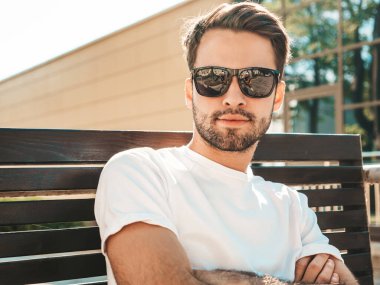Yakışıklı, gülümseyen stil sahibi Lambersex modelinin portresi. Beyaz tişört giyen adam. Moda erkeği, arka planda, güneş gözlüklü bankta oturuyor. Çapraz kollar