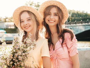 İki genç, güzel, gülümseyen yeniyetme kadın, modaya uygun bir yaz elbisesi içinde. Sokakta şapkalarla poz veren kaygısız seksi kadınlar. Sunset 'te olumlu modeller var. Çiçek tutuyorlar.