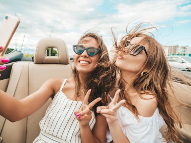 Üstü açık arabadaki iki güzel ve gülümseyen hippi kadının portresi. Cabriolet kullanan kaygısız seksi kadınlar. Pozitif modeller dışarıda güneş gözlüğü takıp eğleniyorlar. Selfie çekiyorlar.