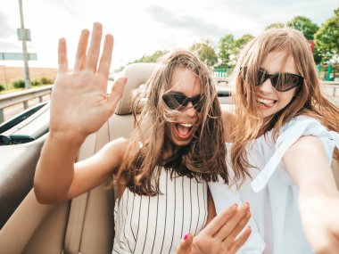 Üstü açık arabadaki iki güzel ve gülümseyen hippi kadının portresi. Cabriolet kullanan kaygısız seksi kadınlar. Pozitif modeller dışarıda güneş gözlüğü takıp eğleniyorlar. Yaz günlerinin tadını çıkarıyorlar.