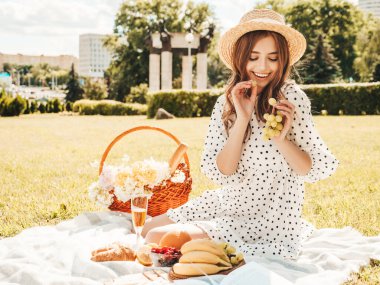 Moda elbisesi ve şapkası olan genç ve güzel hippi kadın. Kaygısız kadın dışarıda piknik yapıyor. Pozitif model çimlerin üzerinde oturuyor, meyve ve peynir yiyor, şampanya içiyor.