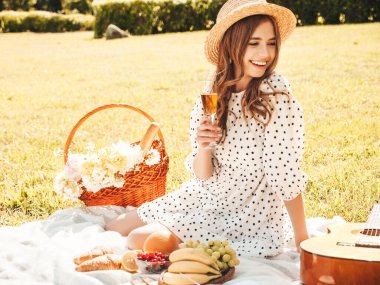 Moda elbisesi ve şapkası olan genç ve güzel hippi kadın. Kaygısız kadın dışarıda piknik yapıyor. Pozitif model çimlerin üzerinde oturuyor, meyve ve peynir yiyor, şampanya içiyor.